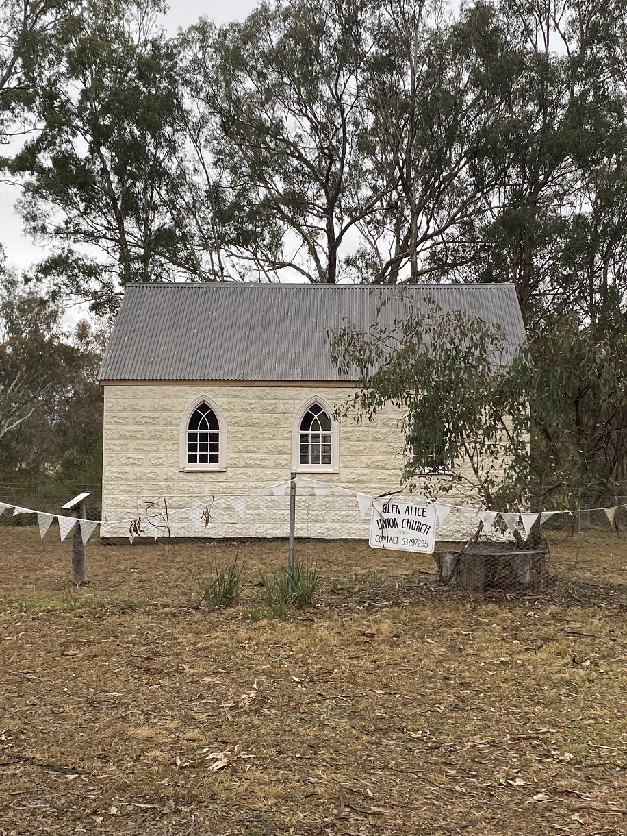 White Glen Alice church building nestled in the trees and featuring arch type windows. In the foreground is a fence with church banner and tree.