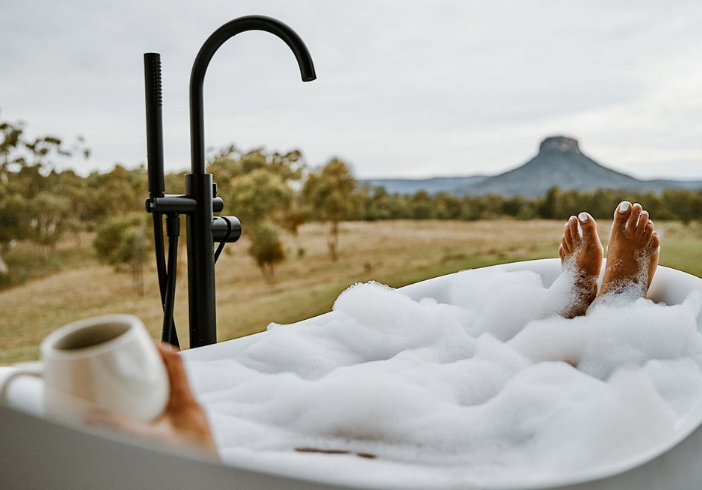 A person lazing in a foamy bath with a cuppa, looking at the view of Pantoneys Crown, in the Capertee Valley, NSW. There is a tap faucet between the bath and the view.